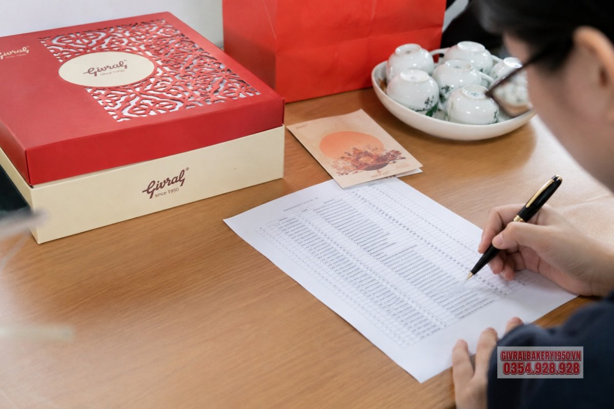 Một bàn làm việc văn phòng với danh sách quà tặng trung thu, hộp bánh Givral sang trọng, thiệp chúc mừng và bút ký, thể hiện việc chuẩn bị quà cho nhân viên hoặc đối tác.